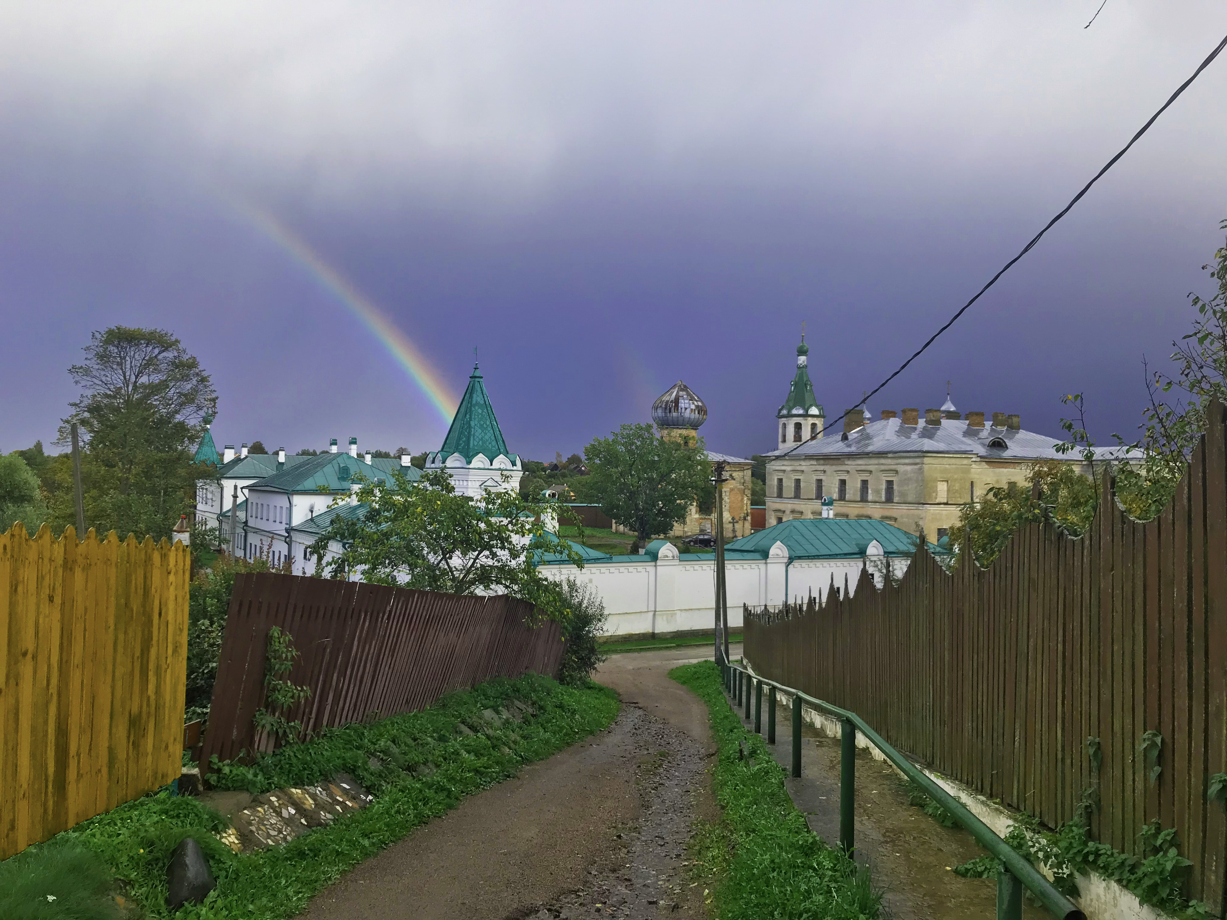 A small dirt alley situated between two wooden fences looks out towards the green and white St. Nicholas monastery. The sky is stormy and a rainbow peeks through the gray.