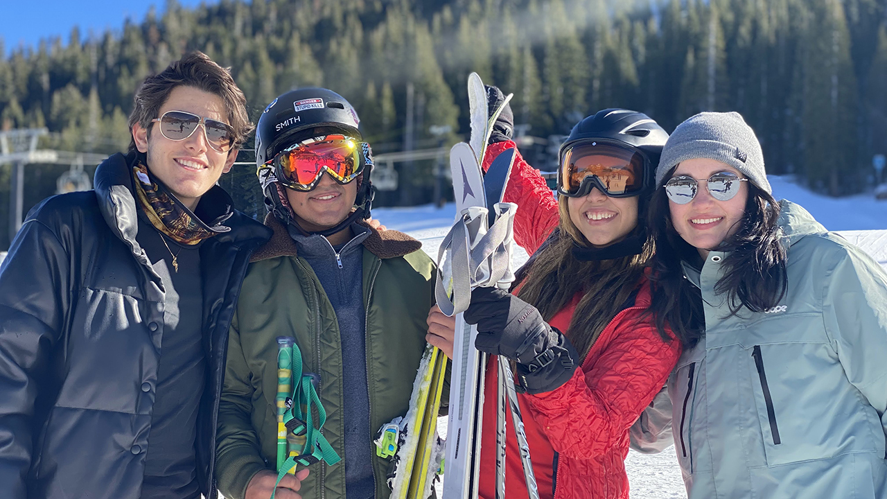 Four students in the snow. The student in the middle of the frame is holding skis.