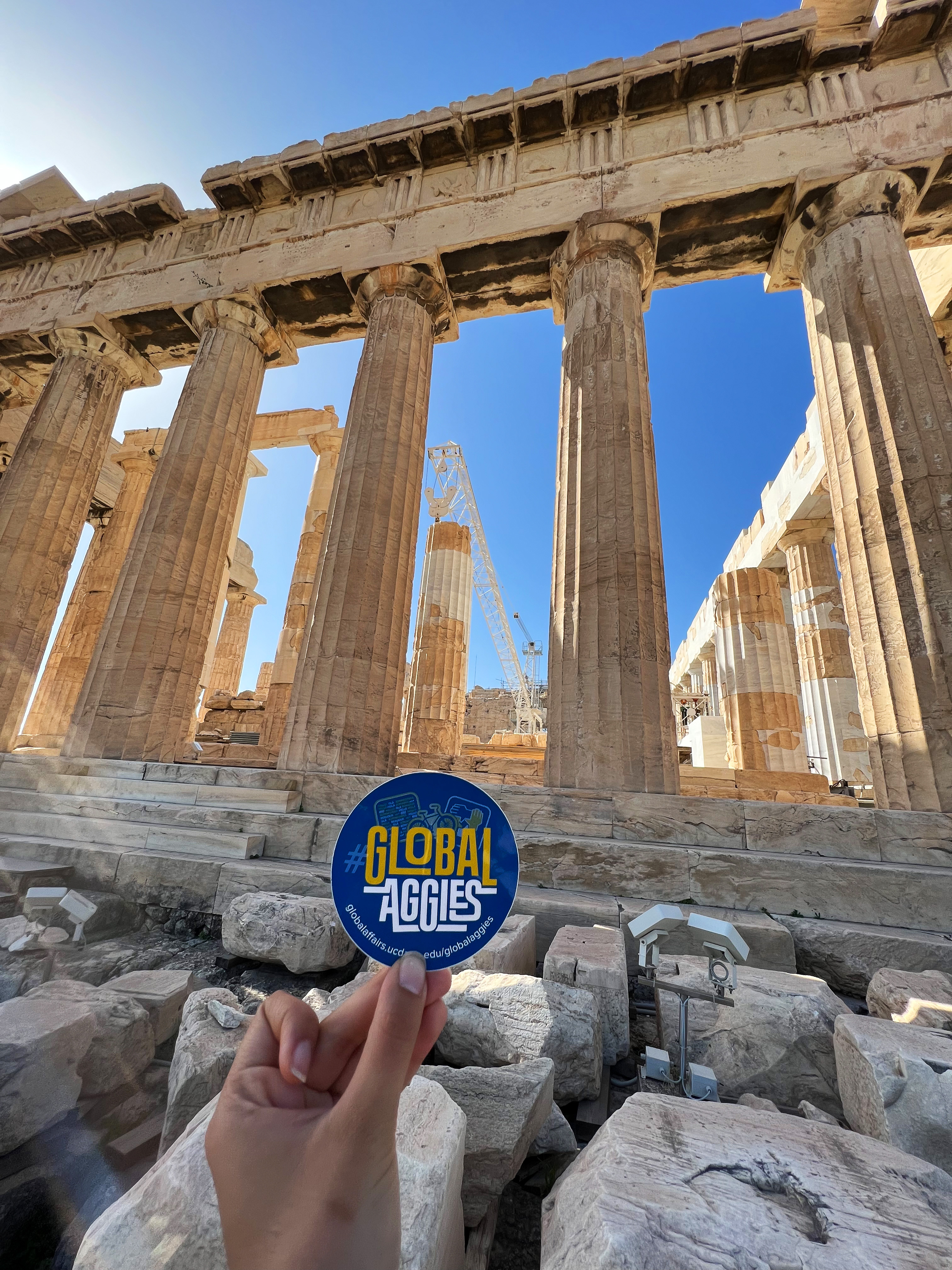 Student holding up a Global Aggies sticker in front of the Parthenon in Greece.