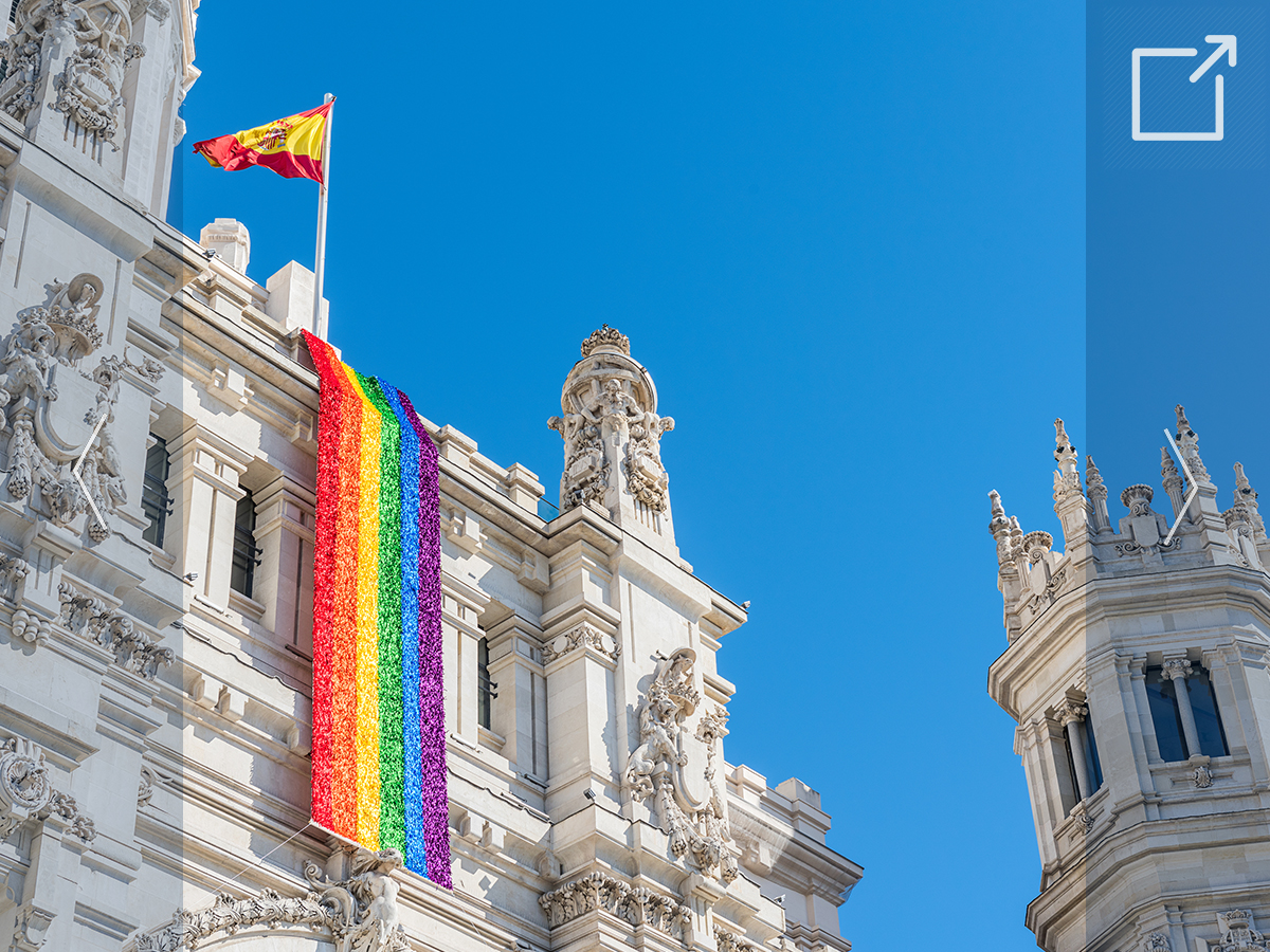 Image of a large building in Madrid, Spain, with a Spanish flag at the top and a long rainbow pride flag handing down the center