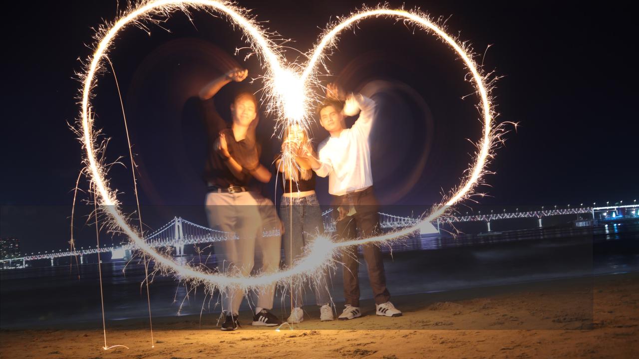 Photo of three students on a beach at night. They are waving hand fire works in the shape of a heart.