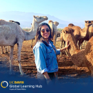 Photo of a student standing next to camels in Morocco. Her left hand is petting the neck of a large camel.