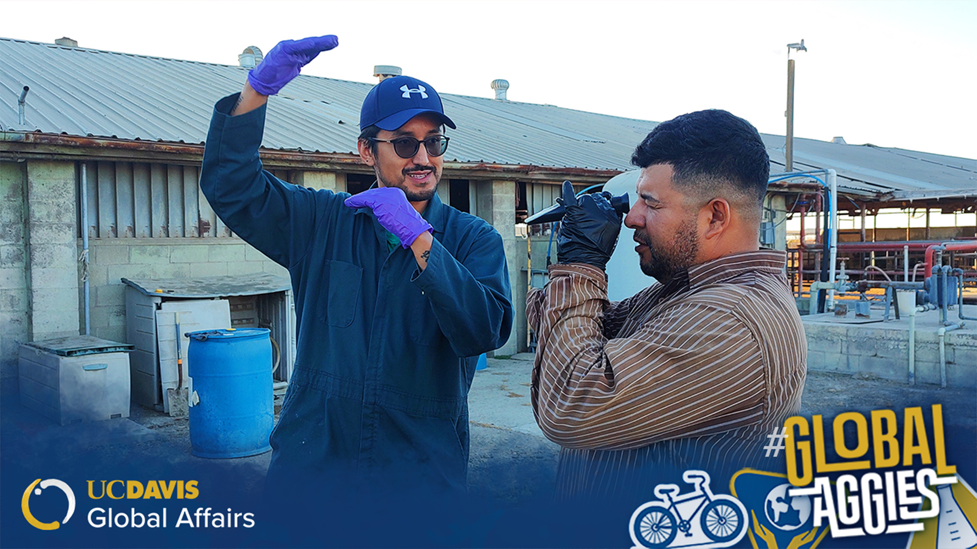 The image depicts two individuals standing outdoors in front of a building with a corrugated metal roof, on what appears to be an industrial or farm site. One person is wearing a blue baseball cap, sunglasses, a dark blue jumpsuit, and purple gloves, gesturing with hands making a frame-like shape. The second person is dressed in a striped long-sleeve shirt, wearing black gloves, and is looking through a handheld device, possibly a refractometer. In the background, there are large blue plastic barrels and ut