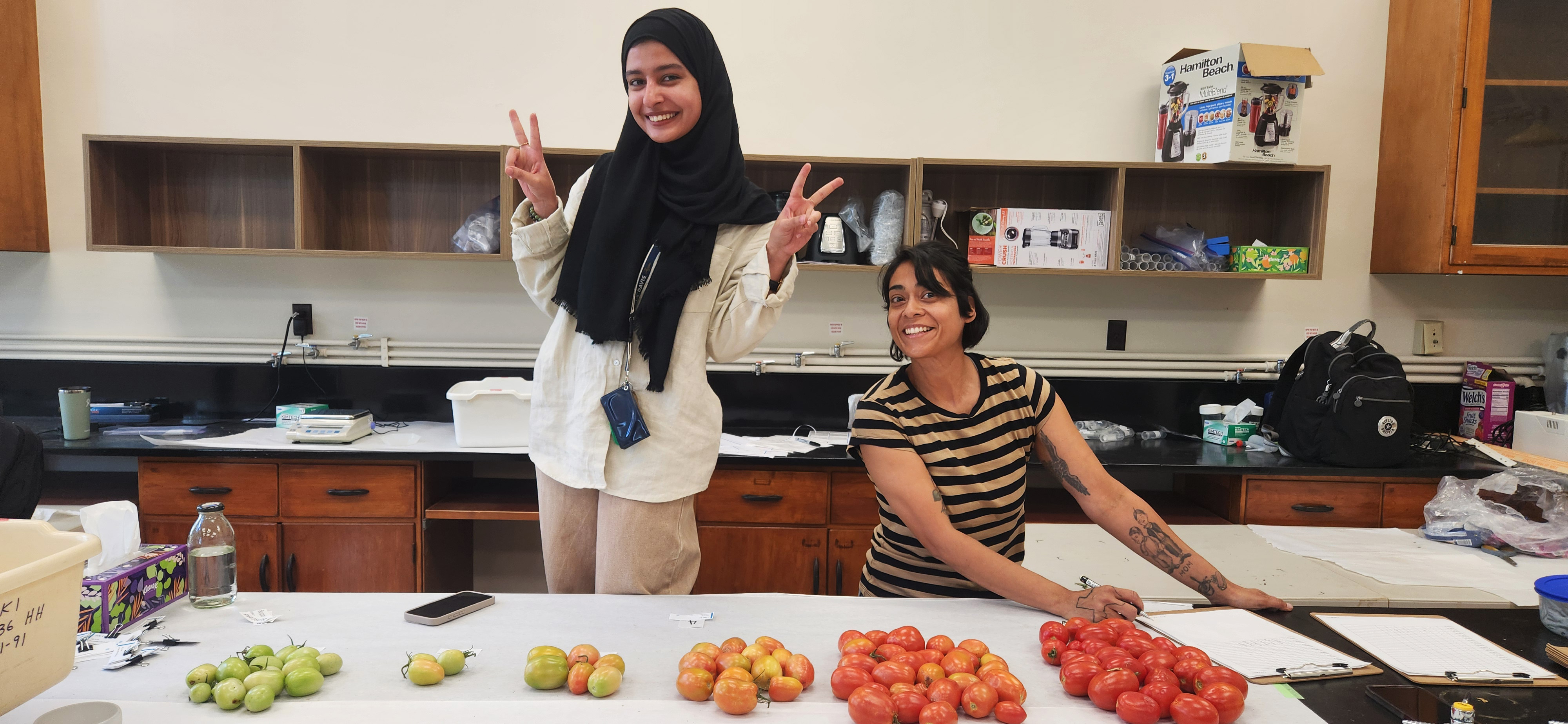 Two women at a table displaying rows of tomatoes organized by ripeness. One, standing, wears a black hijab and beige clothes, showing peace signs. The other, leaning on the table, wears a striped shirt and smiles widely. Shelves, a backpack, and equipment are in the background.