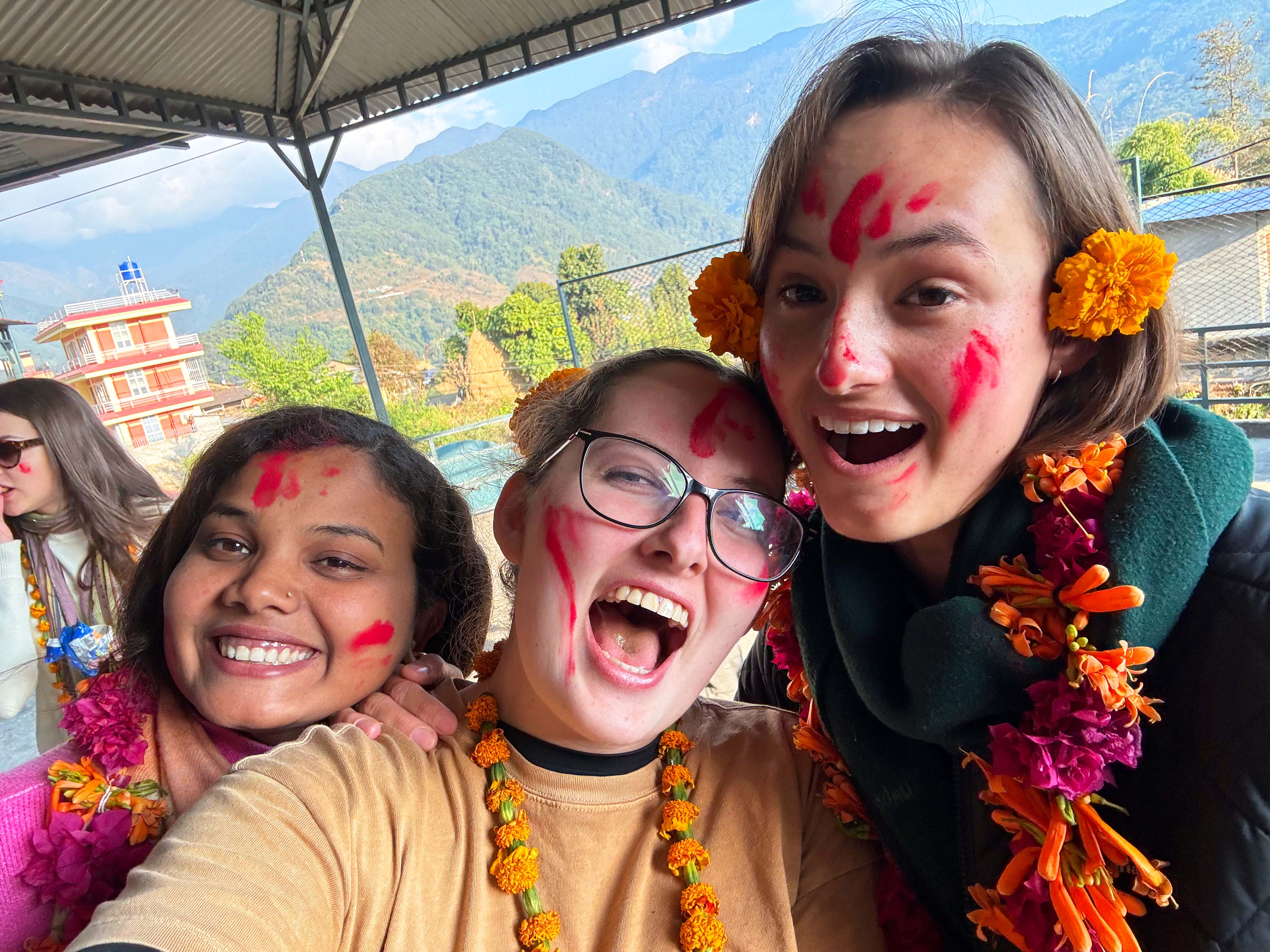 [Three smiling individuals with colorful garlands of orange and pink flowers around their necks pose for a selfie. Their faces are adorned with red powder, suggesting they participated in a cultural celebration. Behind them, a building and lush green mountains are visible, partially obscured by the roof of the shelter they are under. The scene is vibrant and joyful.