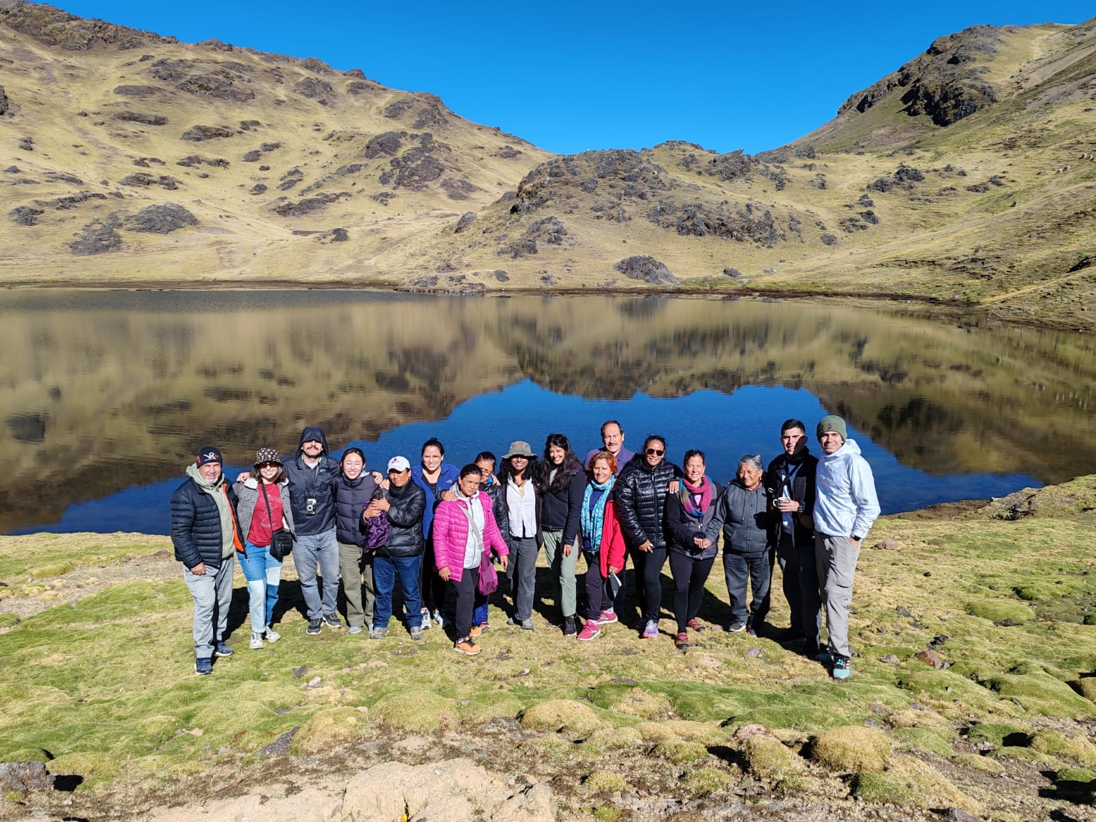 A group of people stand together in front of a serene mountain lake with clear blue skies and rolling hills reflected in the still water. The group is dressed in outdoor gear, including jackets and hats, standing on grassy terrain. The rocky hills and green landscape surround the lake, creating a peaceful, natural setting. The group members are smiling, posing for the photo in this remote, scenic location.