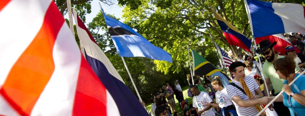 A lively outdoor scene with people holding various national flags during a multicultural event. The flags, including those of the United States, France, Venezuela, Tanzania, and others, are prominently displayed. The crowd is diverse, with people of different ages and backgrounds engaging in conversation. The setting is shaded by trees, creating a festive and inclusive atmosphere.