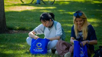 Two students seated on the grass, each with UC Davis Study Abroad bags beside them, enjoying a sunny day outdoors.
