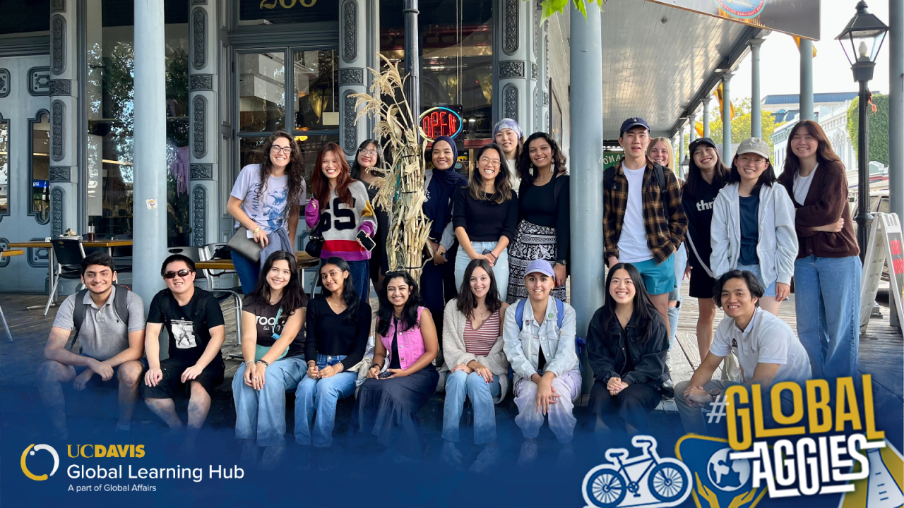 A diverse group of students poses for a photo outside a café with ornate blue-gray doors. Some are sitting while others stand behind them, all smiling. The image includes a UC Davis Global Learning Hub logo and a #GlobalAggies graphic overlay.