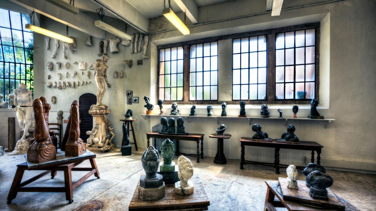 A spacious artist's studio filled with various sculptures and busts. The room has large, grid-like windows letting in natural light. Numerous small busts are arranged on a long shelf along the wall, while a variety of sculpted body parts, including arms and legs, hang on the wall as decoration. The center of the room features several stone and marble sculptures on wooden tables, including large carved figures and intricate designs.
