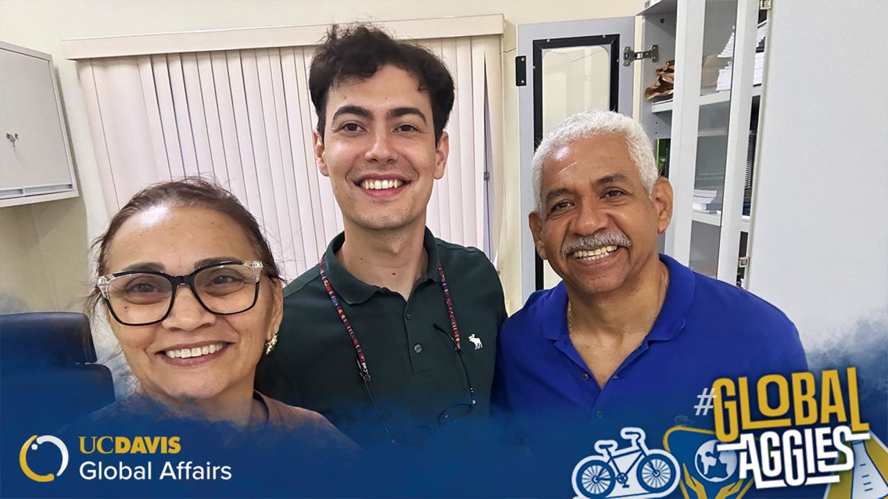 Three people smiling and posing for a selfie indoors, with a backdrop of office furniture and blinds. The group includes a woman wearing glasses, a young man with curly hair, and an older man with gray hair and a mustache. They appear happy and engaged. The image is branded with 'UC Davis Global Affairs' and 'Global Aggies' logos at the bottom corner.