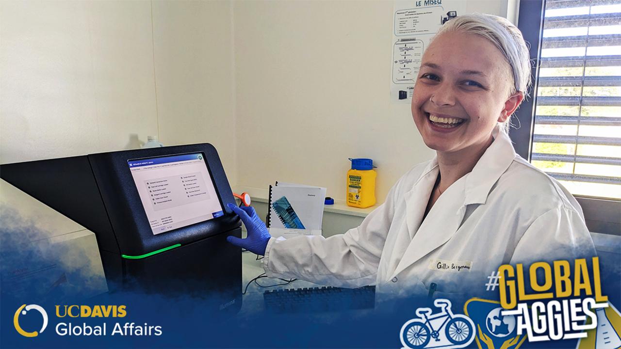 The image features a person wearing a white lab coat and blue gloves, smiling broadly at the camera. They are in a laboratory setting, standing beside a black monitor displaying a screen with various options. The room has cream-colored walls and a poster on the wall behind them. Next to the monitor, there is a yellow biohazard disposal container. A gray window with horizontal blinds allows light into the room. At the bottom left, there is a blue and gold overlay with the UC Davis Global Affairs logo...