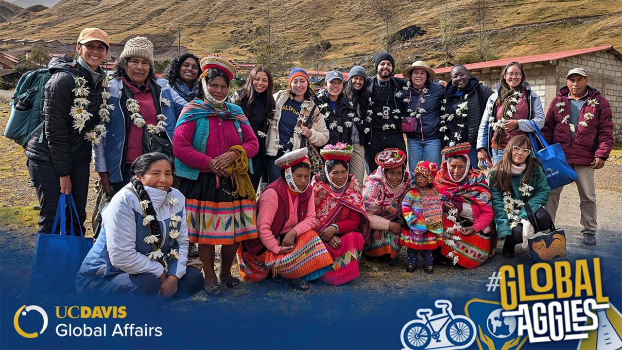 A group of UC Davis students and local community members in traditional clothing pose together outdoors in a mountainous rural area. Some people are wearing garlands made of white flowers. The women in the front row are dressed in vibrant woven garments, with colorful skirts and headwear. The students stand behind them, smiling, many dressed in casual outdoor gear. In the bottom left corner, the UC Davis Global Affairs logo is visible, and in the bottom right, the hashtag '#GlobalAggies' is displayed.