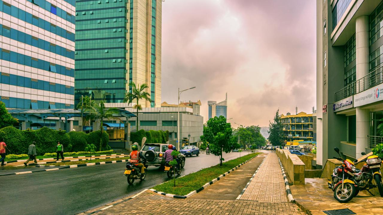 A city street scene in Kigali, Rwanda, featuring modern high-rise buildings with reflective glass facades on both sides. Motorcyclists and a few pedestrians navigate the clean, well-maintained road bordered by manicured hedges and sidewalks. The sky is overcast with gray clouds, adding a dramatic backdrop to the urban landscape. In the distance, more buildings, including a distinctively shaped skyscraper, contribute to the city's skyline.