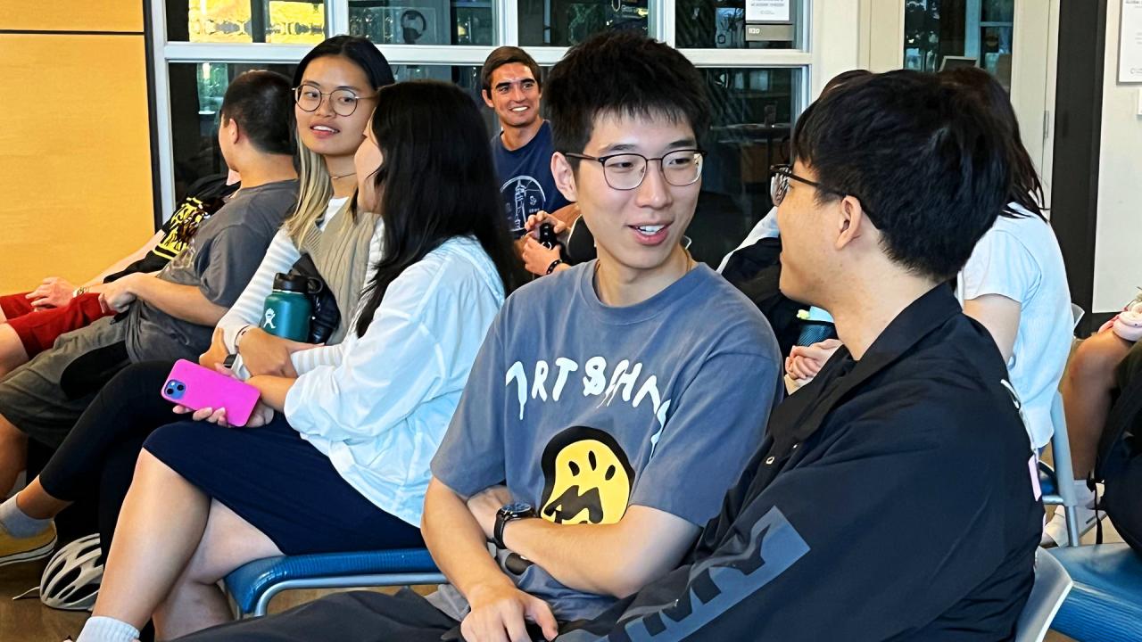 A group of students sitting in a casual setting, engaging in conversation. Two students in the foreground are facing each other, smiling as they talk. The student on the left is wearing glasses and a gray graphic T-shirt with a yellow face design, while the student on the right is also wearing glasses and a dark jacket. In the background, other students are seated, some holding phones or water bottles, with one student looking towards the camera and smiling.