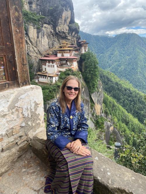 A woman wearing traditional Bhutanese attire sits on a stone ledge with a serene smile. Behind her, the famous Tiger’s Nest Monastery is nestled into a cliffside, surrounded by lush green mountains under a partly cloudy sky.