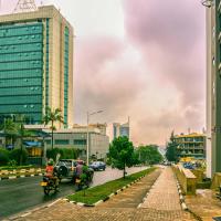 A city street scene in Kigali, Rwanda, featuring modern high-rise buildings with reflective glass facades on both sides. Motorcyclists and a few pedestrians navigate the clean, well-maintained road bordered by manicured hedges and sidewalks. The sky is overcast with gray clouds, adding a dramatic backdrop to the urban landscape. In the distance, more buildings, including a distinctively shaped skyscraper, contribute to the city's skyline.
