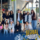 A diverse group of students poses for a photo outside a café with ornate blue-gray doors. Some are sitting while others stand behind them, all smiling. The image includes a UC Davis Global Learning Hub logo and a #GlobalAggies graphic overlay.