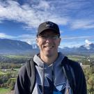Scott smiles in a black San Jose Sharks baseball cap and sweatshirt in front of a scenic view with mountains in the background.