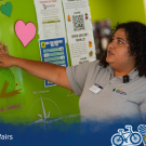 A student volunteer in a gray polo points to a QR code posted on a board at the Aggie Compass. A blue overlay with the Global Affairs logo in the left corner and Global Aggies in the right sits along the bottom of the image.