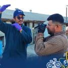 The image depicts two individuals standing outdoors in front of a building with a corrugated metal roof, on what appears to be an industrial or farm site. One person is wearing a blue baseball cap, sunglasses, a dark blue jumpsuit, and purple gloves, gesturing with hands making a frame-like shape. The second person is dressed in a striped long-sleeve shirt, wearing black gloves, and is looking through a handheld device, possibly a refractometer. In the background, there are large blue plastic barrels and ut