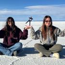 On a sunlit salt flat, two women wearing sunglasses sit on the ground, playfully holding up smaller figures in their hands through forced perspective. The smaller figures in the background stand in yoga poses, with the salt flats stretching out beneath a wide, clear sky.