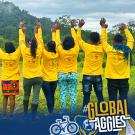 Group of ten people standing outdoors with their backs to the camera, all wearing bright yellow long-sleeve shirts and raising their arms in celebration. They are surrounded by lush green hills and trees under a cloudy sky. The bottom of the image displays the UC Davis Global Affairs logo and the hashtag '#GlobalAggies' with icons of a bicycle and a globe.