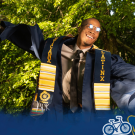 Mahiri Comeaux leaps in the air with arms outstretched wearing a commencement robe and Chicanx/Latinx stole