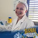 The image features a person wearing a white lab coat and blue gloves, smiling broadly at the camera. They are in a laboratory setting, standing beside a black monitor displaying a screen with various options. The room has cream-colored walls and a poster on the wall behind them. Next to the monitor, there is a yellow biohazard disposal container. A gray window with horizontal blinds allows light into the room. At the bottom left, there is a blue and gold overlay with the UC Davis Global Affairs logo...