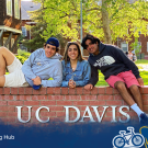 Three students sitting on a large, brink wall. The wall has large text: UC Davis. There is a graphic overlay that reads: Aggies Abroad.