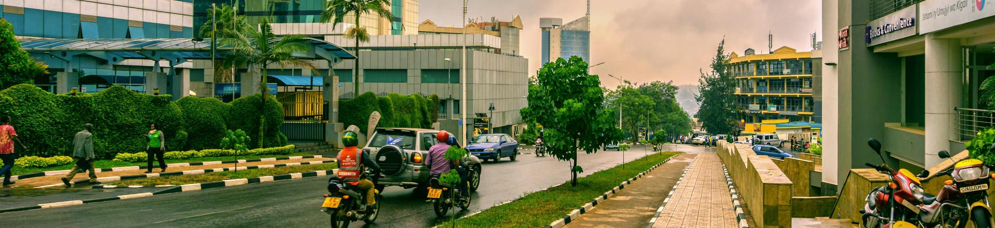 A city street scene in Kigali, Rwanda, featuring modern high-rise buildings with reflective glass facades on both sides. Motorcyclists and a few pedestrians navigate the clean, well-maintained road bordered by manicured hedges and sidewalks. The sky is overcast with gray clouds, adding a dramatic backdrop to the urban landscape. In the distance, more buildings, including a distinctively shaped skyscraper, contribute to the city's skyline.