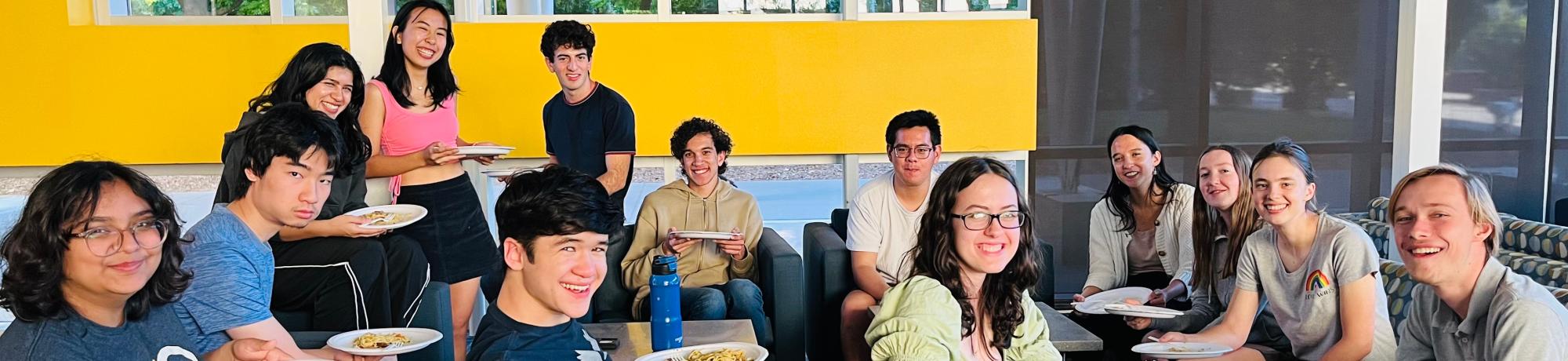 A group students sitting and standing in a brightly lit room, enjoying a meal together.