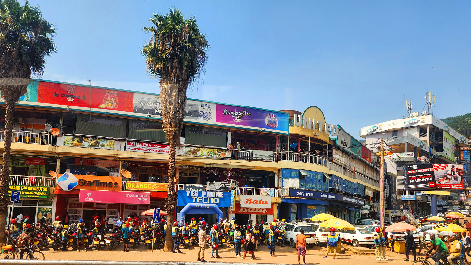 Busy street in Rwanda with colorful storefronts, palm trees, motorcycles, and crowds of people under a clear blue sky.
