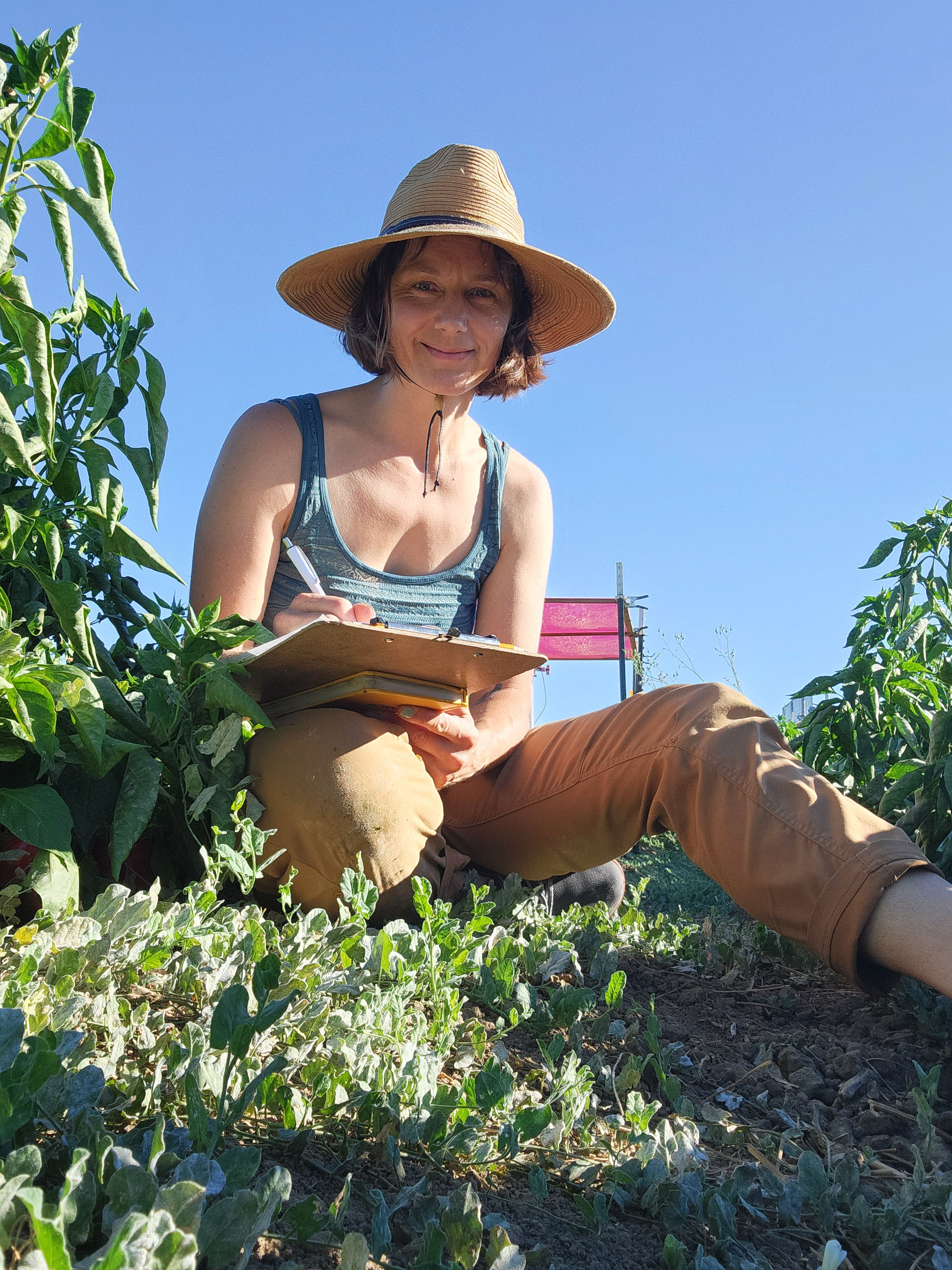 A woman wearing a straw hat, a tank top, and tan pants sits on the ground in a garden. She holds a clipboard and pen, smiling while surrounded by green plants under a bright blue sky.