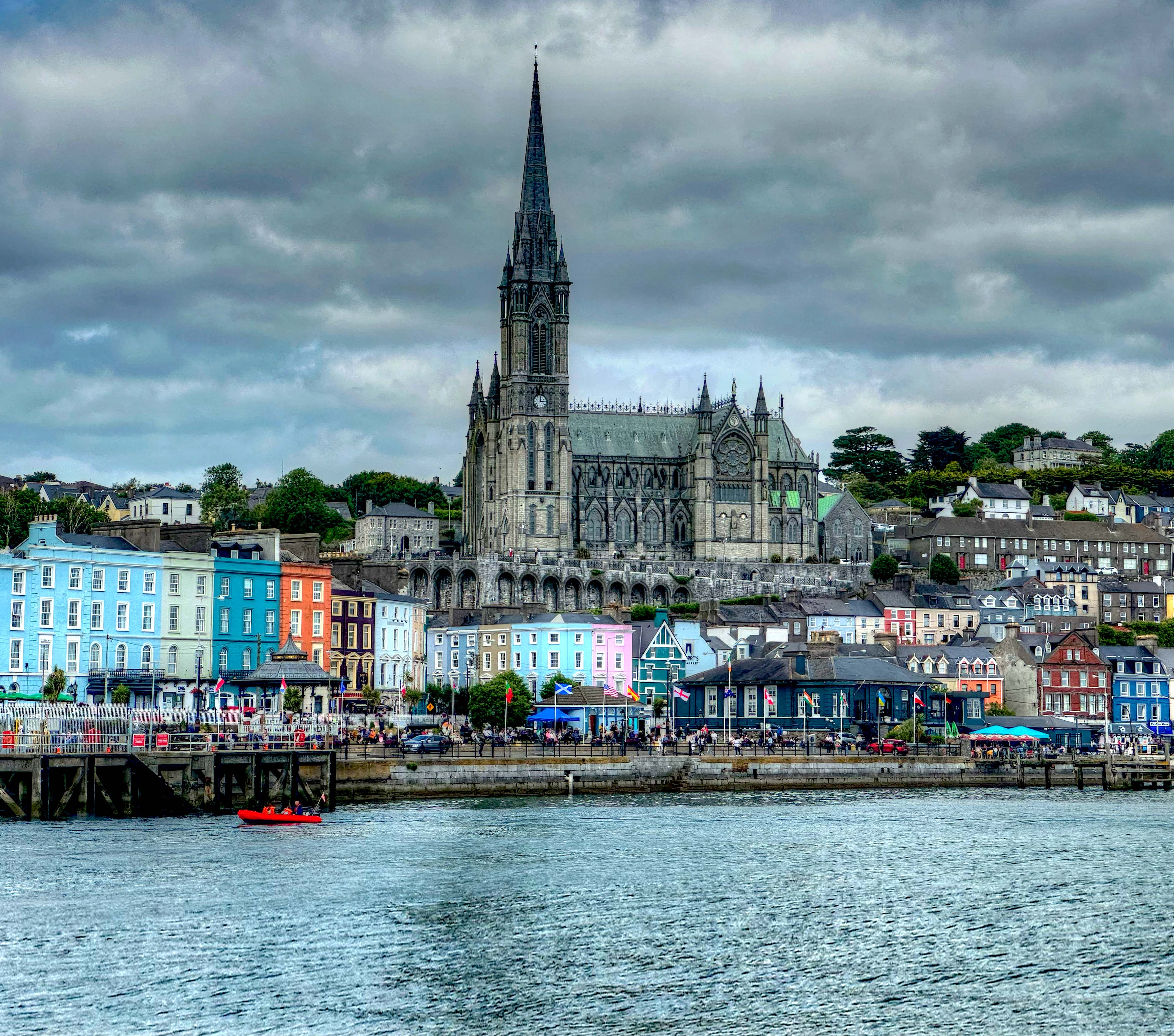 A waterfront view of Cobh, Ireland, featuring St. Colman’s Cathedral with its tall Gothic spire rising above rows of colorful buildings. The town's houses are painted in bright hues of blue, yellow, and pink, contrasting with the overcast sky. A small red boat floats in the foreground on calm water.