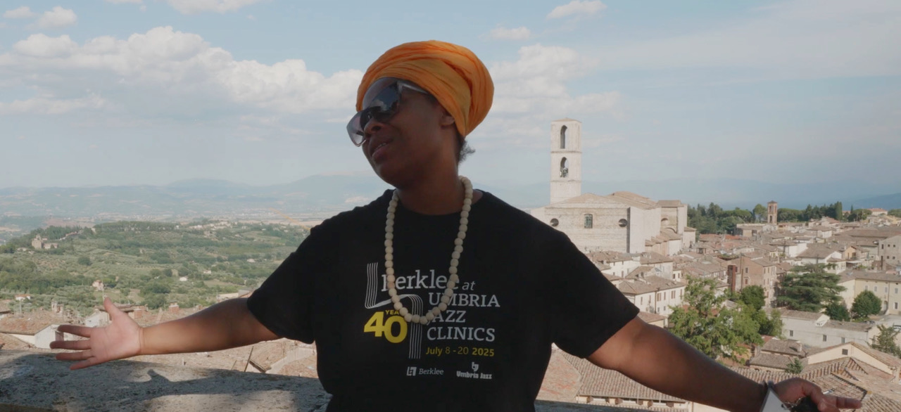 Person in orange headwrap and sunglasses, arms outstretched over sunlit hill town