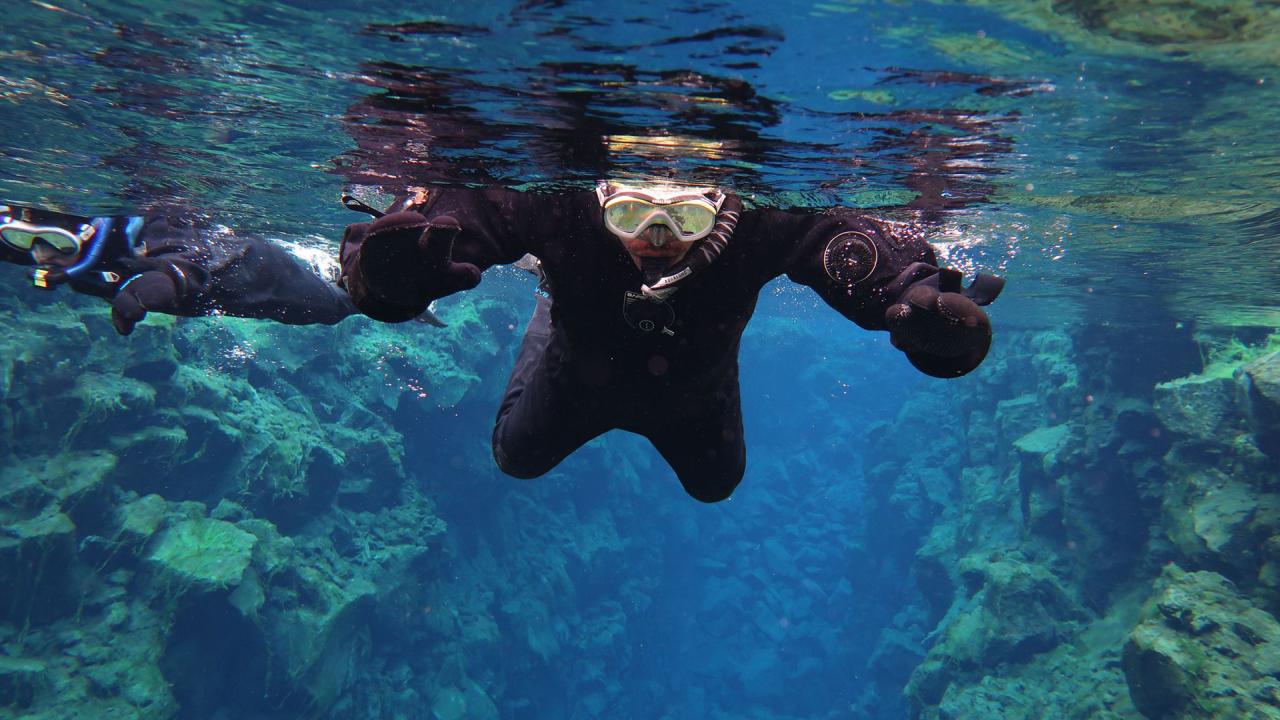 Two snorkelers in black drysuits swim in clear blue water above rocky underwater formations.