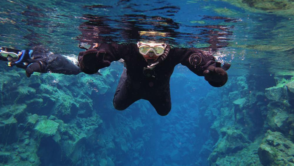 Two snorkelers in black drysuits swim in clear blue water above rocky underwater formations.