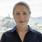 A woman with light hair and a navy shirt smiles, with a city and water background.