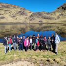 A group of people stand together in front of a serene mountain lake with clear blue skies and rolling hills reflected in the still water. The group is dressed in outdoor gear, including jackets and hats, standing on grassy terrain. The rocky hills and green landscape surround the lake, creating a peaceful, natural setting. The group members are smiling, posing for the photo in this remote, scenic location.