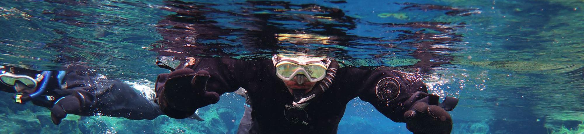 Two snorkelers in black drysuits swim in clear blue water above rocky underwater formations.