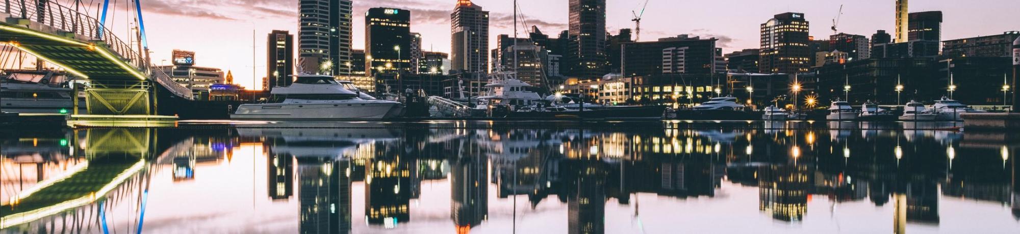 City skyline reflecting on water at dusk, with boats moored in the foreground.