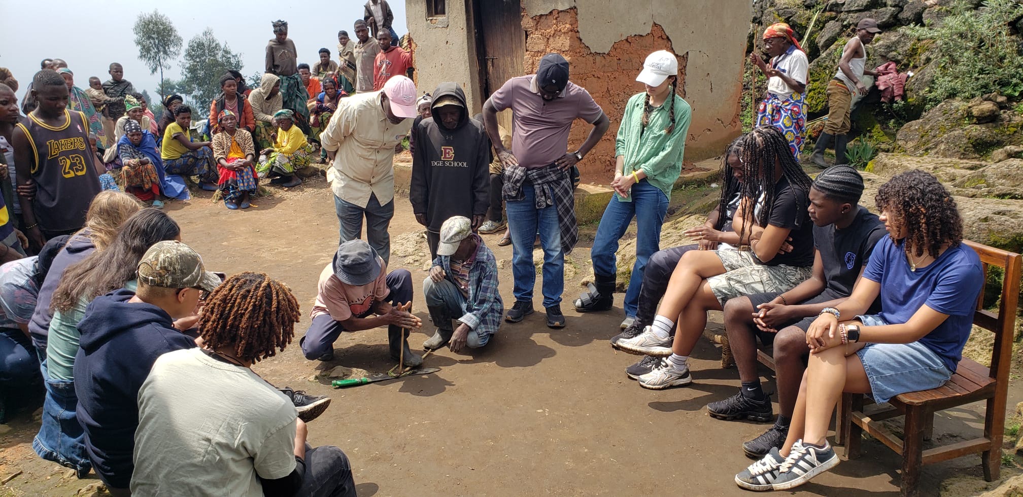 Outdoor community meeting on a dirt path; people seated and standing, listening attentively