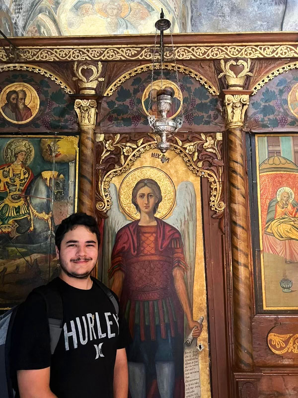 Student standing adjacent to a wall mural depiction of the Archangel. Photo captured at the Archaeological Site of Mystras.