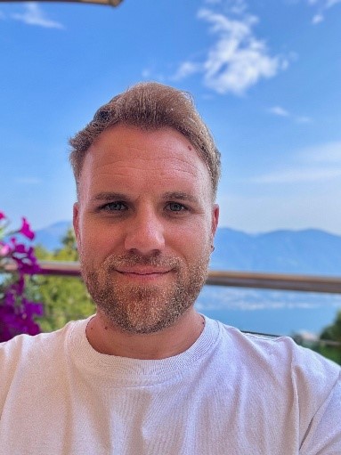Casual headshot photograph of smiling man in white shirt on balcony with mountains