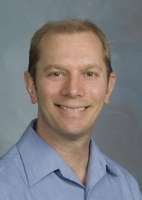 Smiling man with short light brown hair, wearing a blue collared shirt against a gray background.