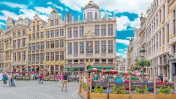 Colorful buildings line a cobblestone square, with outdoor seating and a blue sky above.