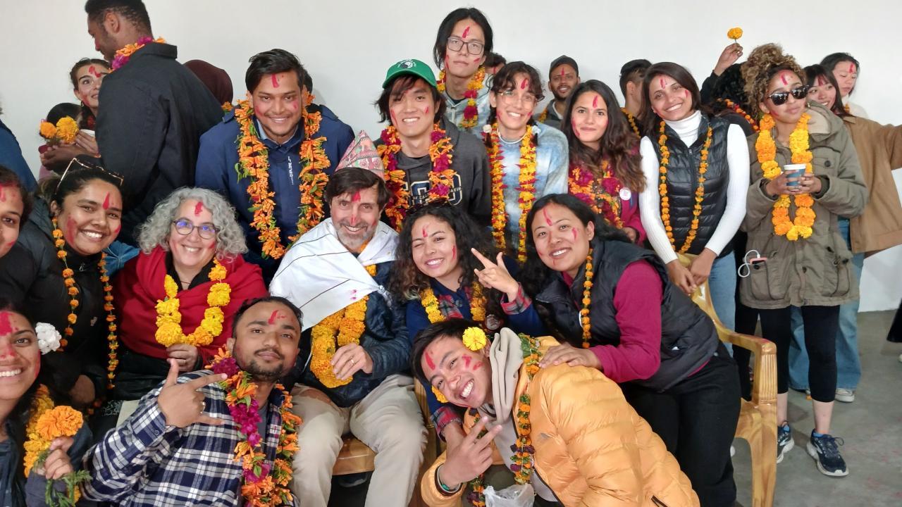 A joyful group of students in Nepal celebrating together, adorned with flower garlands and colored powder.