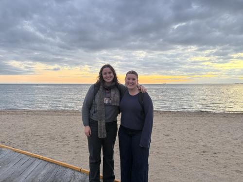 Two women stand on a beach at sunset, with clouds above and water in the background.