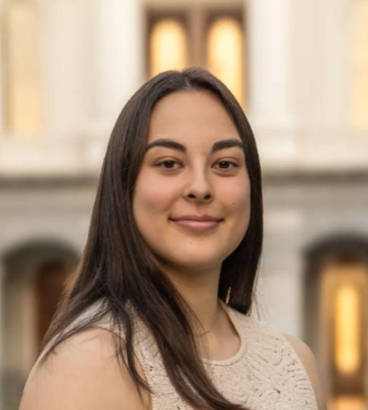 Portrait of young woman with long dark hair in beige top, smiling before blurred columned building